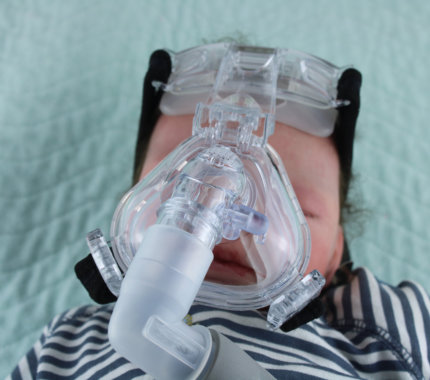 an infant wearing a CPAP mask with headgear, lying on a bed with a light blue quilted blanket in the background.